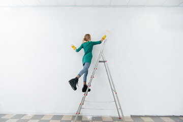 A young girl on a stepladder paints a white wall with a roller. Repair of the interior.