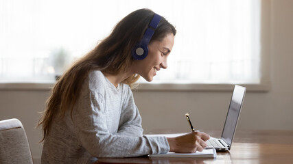 Smiling millennial teen female in headphones look at laptop screen write study online at home....