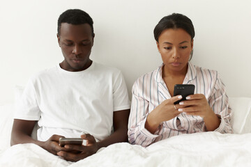 Mixed race couple of freelancers using gadgets for distant work. Serious young black man and woman working from bed, glued to screens of their mobile phones, typing fast. Technology and addiction