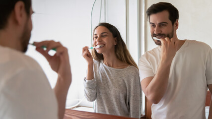 Happy millennial multiracial newlyweds couple look in mirror in new home bathroom brush teeth together. Smiling young man and woman enjoy early morning washing routine in bath. Rental concept.