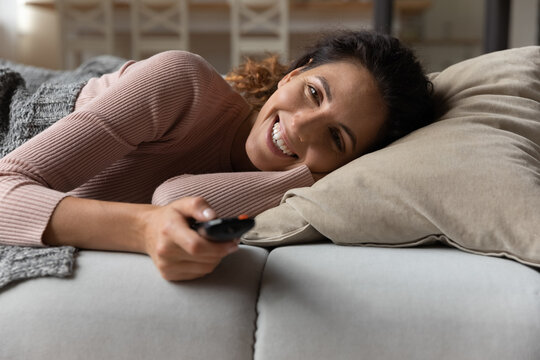 Happy Latina Female Rest On Cozy Couch Lie In Comfortable Pose Covered With Plaid Hold Tv Remote Choose Entertainment Channel. Smiling Young Lady Watch Television At Home Having Fun Enjoy Comedy Movie