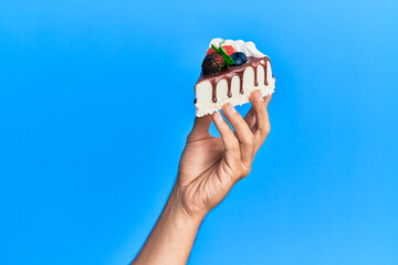 Hand of hispanic man holding slice of chocolate cake over isolated blue background. - Powered by Adobe