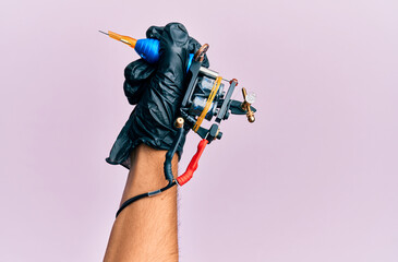 Hand of young hispanic man using tattoo machine over isolated pink background.
