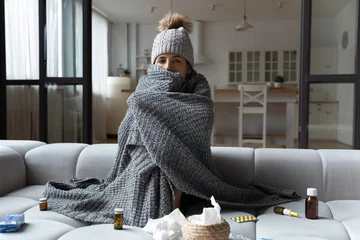 Poster Sick latin female feel bad ill has health problems in cold freezing flat with broken central heating. Young woman sit on sofa in cap covered with knitted plaid taking medicine for fever flu influenza © fizkes