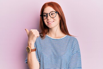 Young red head girl wearing casual clothes and glasses pointing thumb up to the side smiling happy with open mouth