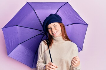 Young red head girl holding purple umbrella wearing fresh beret screaming proud, celebrating victory and success very excited with raised arms