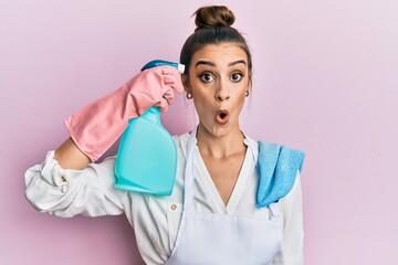 Beautiful brunette young woman wearing apron holding cleaning spray pointing to head scared and...
