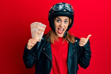 Hispanic young woman wearing motorcycle helmet holding 50 turkish lira pointing thumb up to the side smiling happy with open mouth