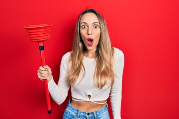 Beautiful hispanic woman holding toilet plunger scared and amazed with open mouth for surprise, disbelief face