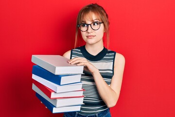 Redhead young woman holding a pile of books smiling looking to the side and staring away thinking.