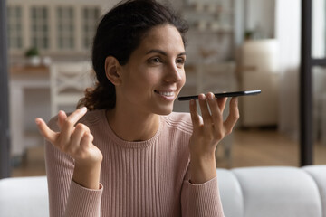 Inspired by innovations. Happy young hispanic woman sit on couch speak to phone ai give audio...
