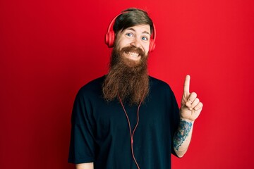 Redhead man with long beard listening to music using headphones with a big smile on face, pointing with hand and finger to the side looking at the camera.