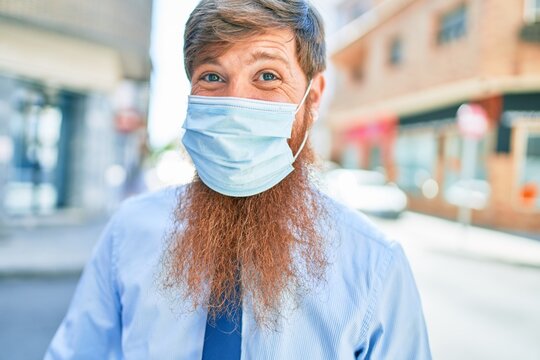 Young handsome redhead businessman wearing coronavirus protection medica mask standing at street of city.
