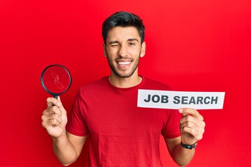 Young handsome man holding magnifying glass for job search winking looking at the camera with sexy...