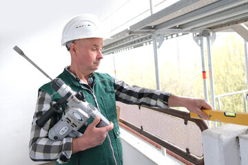 elderly builder with a hand tool in a protective white helmet and working uniform stands at a construction site for the construction and repair of buildings, civil engineering concept