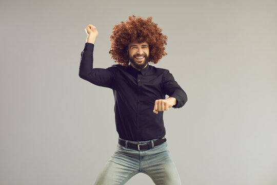 Let's have a party. Happy young man with funny crazy hair having fun in studio. Carefree goofy guy wearing curly wig smiling and dancing gangnam style isolated on gray background