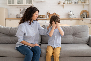 Woman comforting crying son. Disturbed mom touch little upset boy hiding face in hands sitting depressed in couch. Sad kid and stressed young mother together at home. Offended or abused child concept