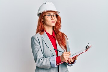 Young redhead woman wearing architect hardhat smiling looking to the side and staring away thinking.