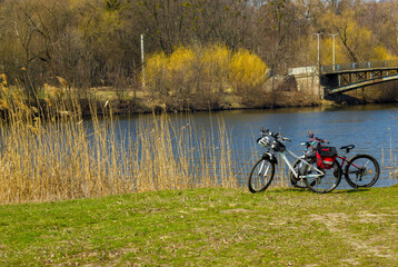 two bicycles by the river in the early spring with bike helmets, gloves, travel bags, young green grass, yellow reeds, old bridge on the background in  a sunny day 