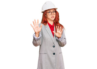 Young redhead woman wearing architect hardhat afraid and terrified with fear expression stop gesture with hands, shouting in shock. panic concept.