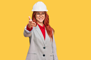 Young redhead woman wearing architect hardhat smiling friendly offering handshake as greeting and welcoming. successful business.