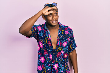 Young black man wearing hawaiian shirt and sunglasses very happy and smiling looking far away with hand over head. searching concept.