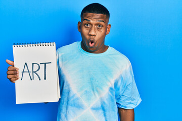 Young black man holding art notebook scared and amazed with open mouth for surprise, disbelief face