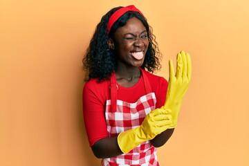 Beautiful african young woman wearing cleaner apron and gloves sticking tongue out happy with funny expression.