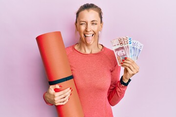 Beautiful caucasian sports woman holding yoga mat and swedish krona banknotes sticking tongue out...
