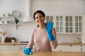 Happy young latin female film video blog for housewives look at camera wave hand in protective glove tidying up kitchen. Portrait of smiling woman vlogger advertise new line of home cleaning products