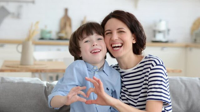 Mom and son making heart hand gesture together happy smiling look in camera at home sitting on sofa. Cute preschool boy and mother take funny family picture photo. Love and happy parenthood concept