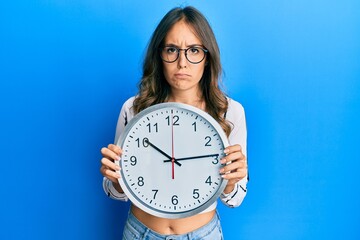 Young brunette woman holding big clock skeptic and nervous, frowning upset because of problem....