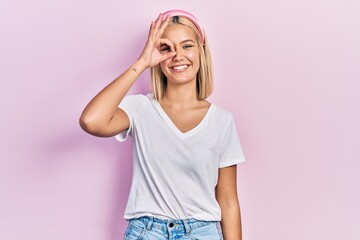 Obraz na płótnie Canvas Beautiful blonde woman wearing casual white t shirt doing ok gesture with hand smiling, eye looking through fingers with happy face.