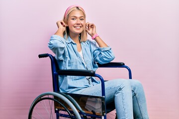 Beautiful blonde woman sitting on wheelchair smiling pulling ears with fingers, funny gesture....