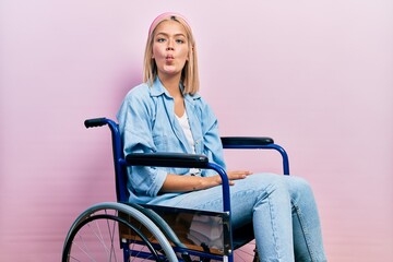 Beautiful blonde woman sitting on wheelchair making fish face with lips, crazy and comical gesture....