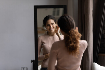 Young beautiful hispanic female stand by big mirror enjoy perfect face skin after home care procedures. Pretty latin woman touch cheek satisfied with good effect after using professional cosmetic line
