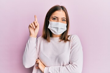 Young beautiful woman wearing medical mask smiling with an idea or question pointing finger up with happy face, number one