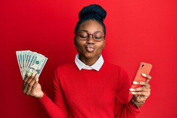 Young african american woman using smartphone holding usa dollars looking at the camera blowing a...