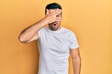 Young hispanic man wearing casual white t shirt peeking in shock covering face and eyes with hand, looking through fingers afraid