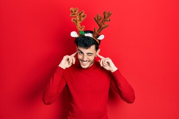 Young hispanic man wearing cute christmas reindeer horns covering ears with fingers with annoyed expression for the noise of loud music. deaf concept.