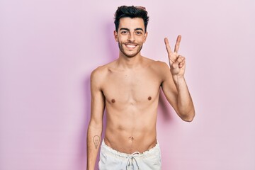 Fototapeta premium Young hispanic man wearing swimwear shirtless showing and pointing up with fingers number two while smiling confident and happy.