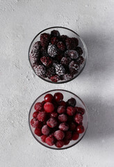 Frozen cherries and blackberries in two glass transparent bowls on gray concrete background. Top view