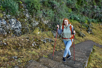 Tourist walking around Fethiye in Turkey, young European woman climbs stairs to Amyntas Rock Tombs using hiking poles.