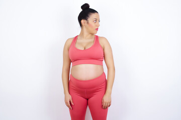 Side view of young happy smiling young beautiful pregnant woman in sports clothes against white wall