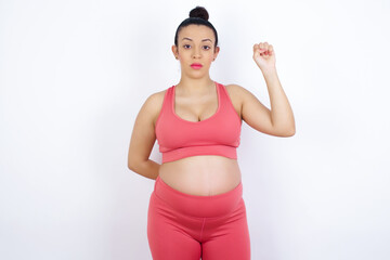 young beautiful Arab pregnant woman in sports clothes against white wall feeling serious, strong and rebellious, raising fist up, protesting or fighting for revolution.