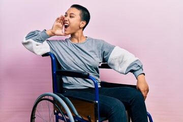 Beautiful hispanic woman with short hair sitting on wheelchair shouting and screaming loud to side with hand on mouth. communication concept.