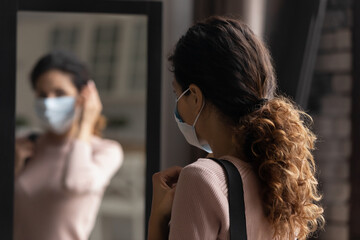 Coronavirus prevention measures. Back view of careful young female putting adjusting single use medical mask on face by big mirror before leaving home to prevent flu covid infection. Selective focus