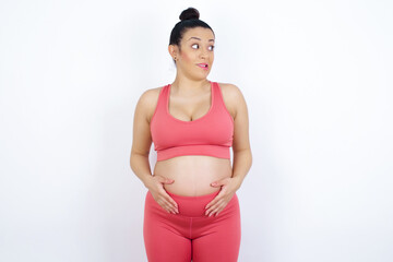 Oops! Portrait of young beautiful Arab pregnant woman in sports clothes against white wall  clenches teeth and looks confusedly aside, realizes her bad mistake,
