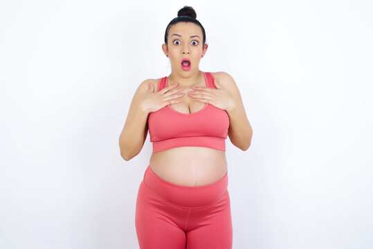 young beautiful Arab pregnant woman in sports clothes against white wall keeps hands on chest feeling shocked and scared, mouth widely opened, stares at camera saying: Who, me?