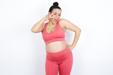 young beautiful Arab pregnant woman in sports clothes against white wall making v-sign near eyes. Leisure, coquettish, celebration, and flirt concept.